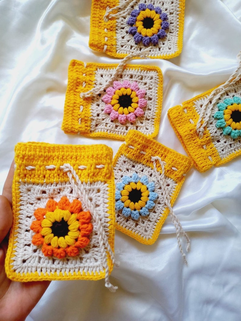 Op de afbeelding: Vijf gehaakte tasjes met zonnebloemmotieven in verschillende kleuren, waaronder geel, roze, blauw en paars. Elk tasje heeft een trekkoord en is gemaakt van wit en geel garen.