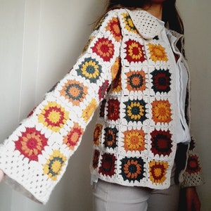 Puede incluir: Una chaqueta de punto de ganchillo blanca con un patrón geométrico colorido. La chaqueta tiene cuello y mangas largas. El patrón está formado por cuadrados con flores de diferentes colores en el centro.