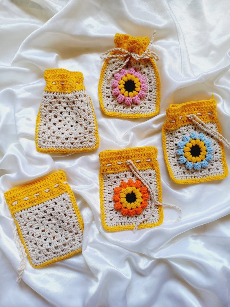 Op de afbeelding: Vijf gehaakte trekkoordtassen in een gele, beige en witte kleurstelling. Elke tas heeft een gehaakt zonnebloemmotief in het midden met bloemblaadjes in verschillende kleuren.