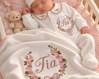 May include: A white long-sleeved baby onesie and matching hat set featuring pink floral embroidery and the name "Tia" in script. A white blanket with matching floral wreath embroidery and the name "Tia" is draped over the baby.