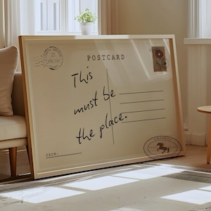 Pode incluir: Grande obra de arte emoldurada que se assemelha a um postal vintage. O texto "This must be the place" está escrito em itálico. A moldura é de madeira clara e o fundo é creme. Selos decorativos são visíveis.