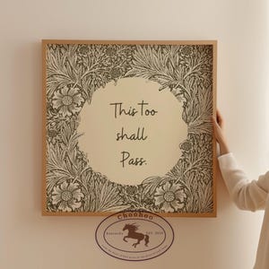 Può includere: Stampa incorniciata con la scritta "This too shall Pass". L'opera presenta un motivo floreale nei toni del verde e del crema, con una cornice in legno marrone chiaro. Un logo circolare con un cavallo è in basso.