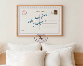 May include: A framed postcard print with a light beige background. The postcard reads "with love from Chicago x" in blue script. It features a postage stamp design and is displayed above white pillows.