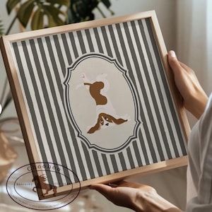 May include: A framed print with a dog illustration. The artwork depicts a brown and white dog in an oval frame set against a background of grey and white vertical stripes. The frame is made of light-coloured wood.
