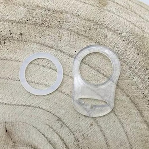 Könnte beinhalten: Zwei transparente Kunststoffringe, einer größer als der andere, auf einer Holzoberfläche. Der größere Ring hat eine flache, rechteckige Form mit einem kleinen Loch in der Mitte. Der kleinere Ring ist rund.