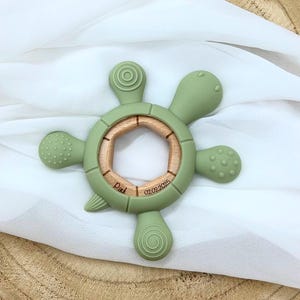 May include: A green silicone baby teether shaped like a turtle with a wooden ring in the center. The teether has textured surfaces and the ring is engraved with the text "Paul 02.02.2025".