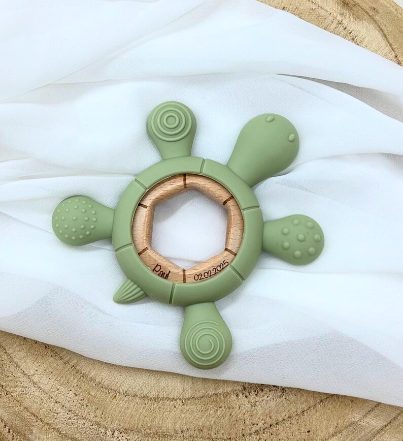 May include: A green silicone baby teether shaped like a turtle with a wooden ring in the center. The teether has textured surfaces and the ring is engraved with the text "Paul 02.02.2025".