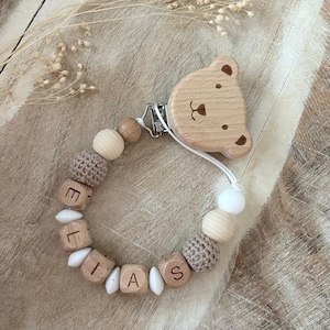 Könnte beinhalten: Ein Schnullerhalter mit einem hellbraunen, bärenförmigen Holzclip an einer Perlenschnur. Die Perlen bestehen aus runden Holzkugeln, beigen gehäkelten Kugeln, weißen rautenförmigen Perlen und Holzwürfeln, die den Namen ELIAS bilden.