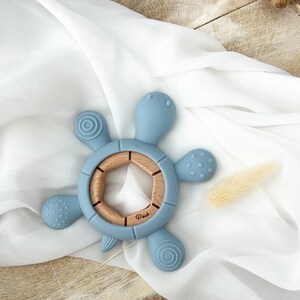 May include: A blue silicone baby teether shaped like a turtle with a wooden center ring. The teether has textured bumps and ridges for baby to explore.