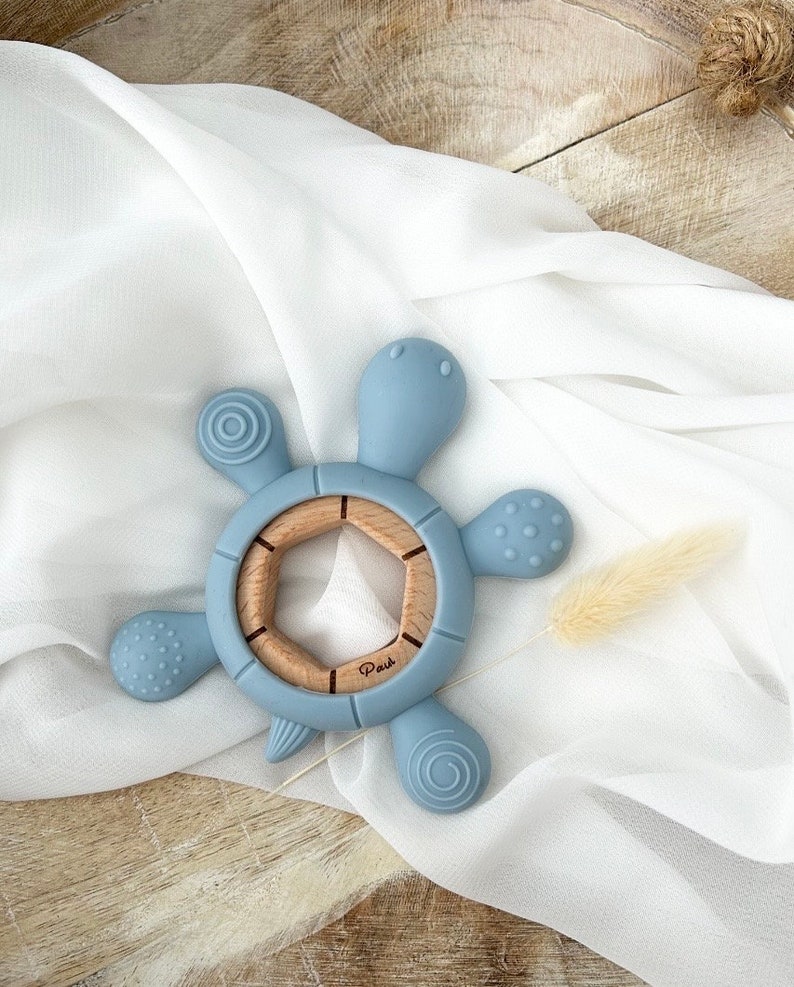 May include: A blue silicone baby teether shaped like a turtle with a wooden center ring. The teether has textured bumps and ridges for baby to explore.