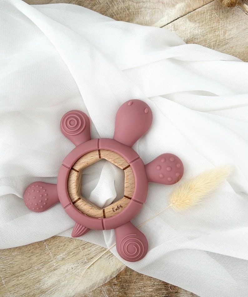 May include: A pink silicone teething toy shaped like a turtle with a wooden ring in the center. The toy has textured surfaces and the word "Emily" is engraved on the wooden ring.