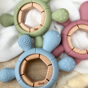 May include: Two silicone teething toys shaped like turtles with wooden centers. The blue turtle has the name "Paul" engraved on the wood. The green turtle has the name "Elias" engraved on the wood.