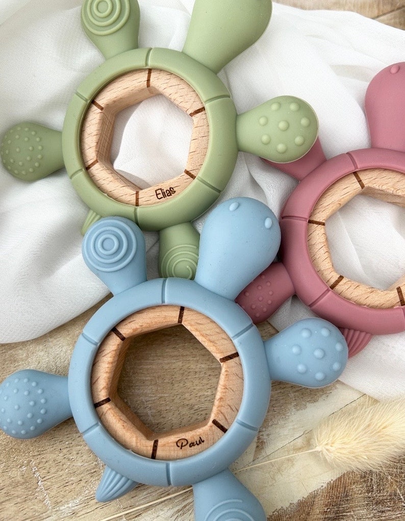 May include: Two silicone teething toys shaped like turtles with wooden centers. The blue turtle has the name "Paul" engraved on the wood. The green turtle has the name "Elias" engraved on the wood.