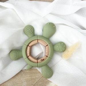 May include: A green silicone turtle teether with a wooden center ring. The teether has textured bumps and ridges for baby's gums. The wooden ring has a hexagonal shape and the word "Elise" is engraved on it.