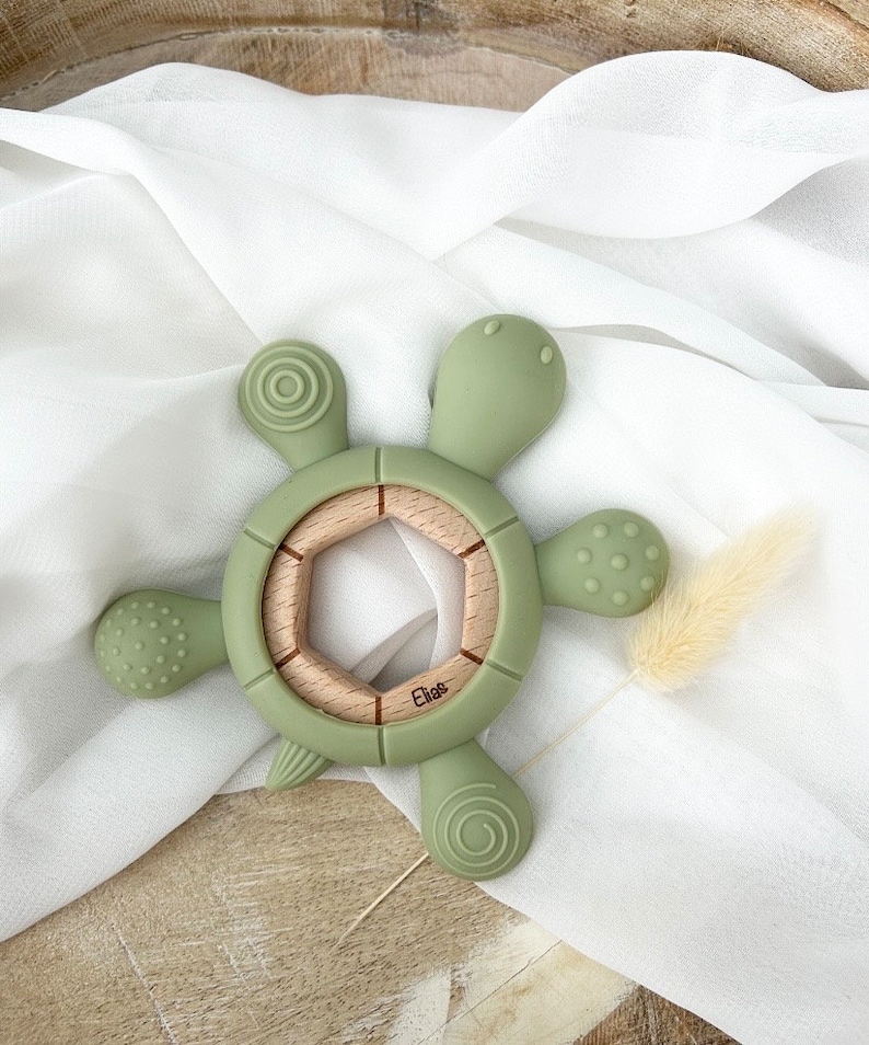 May include: A green silicone turtle teether with a wooden center ring. The teether has textured bumps and ridges for baby's gums. The wooden ring has a hexagonal shape and the word "Elise" is engraved on it.