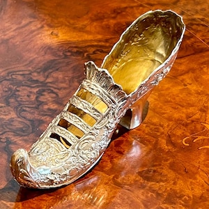 May include: A silver shoe-shaped decorative object with intricate floral designs and openwork details. The shoe is made of metal and has a high heel.