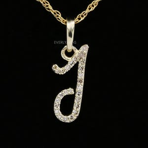 Peut inclure: Un collier pendentif doré avec la lettre "J" en écriture cursive. La lettre est sertie de petites pierres précieuses transparentes. La chaîne du collier est également dorée, et le fond est noir. Le pendentif mesure environ 2,5 cm de haut.