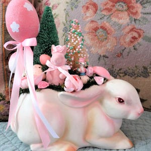Op de afbeelding: Een roze keramische konijnenpot met een roze en witte ballon, roze en groene bomen en roze bloemen. De pot is gevuld met mos en kleine roze eieren.