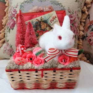 Op de afbeelding: Een wit konijn met rode ogen zit in een rieten mand met een rood-wit gestreept lint. De mand is gevuld met roze bloemen, groen mos en rood-witte paaseieren. Een vintage paaskaarten met de tekst "Easter Greetings" is zichtbaar op de achtergrond.