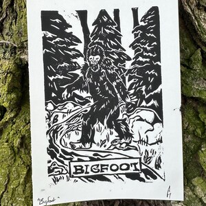 May include: Black and white linocut print of Bigfoot walking through a forest. The print features a sign that reads "BIGFOOT" and is signed "Bigfoot" in the bottom left corner.