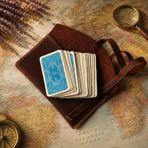 May include: A deck of playing cards with blue patterned backs fanned out on top of a dark brown leather pouch. The cards feature a central illustration of a figure in a frame. The pouch has a wrap-around leather strap and visible stitching.