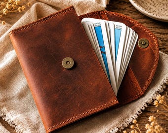 May include: A brown leather tarot card holder, open to reveal a deck of cards with blue and white designs. The holder has a magnetic snap closure and is set on a beige cloth.