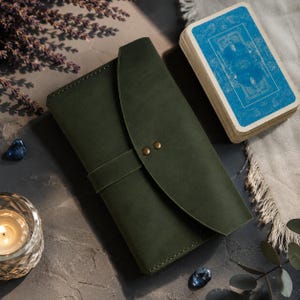May include: A dark olive green leather journal cover featuring a curved flap closure secured by two small brass-colored metal rivets. The rectangular cover has visible stitching along the edges and a horizontal strap that holds the flap in place.