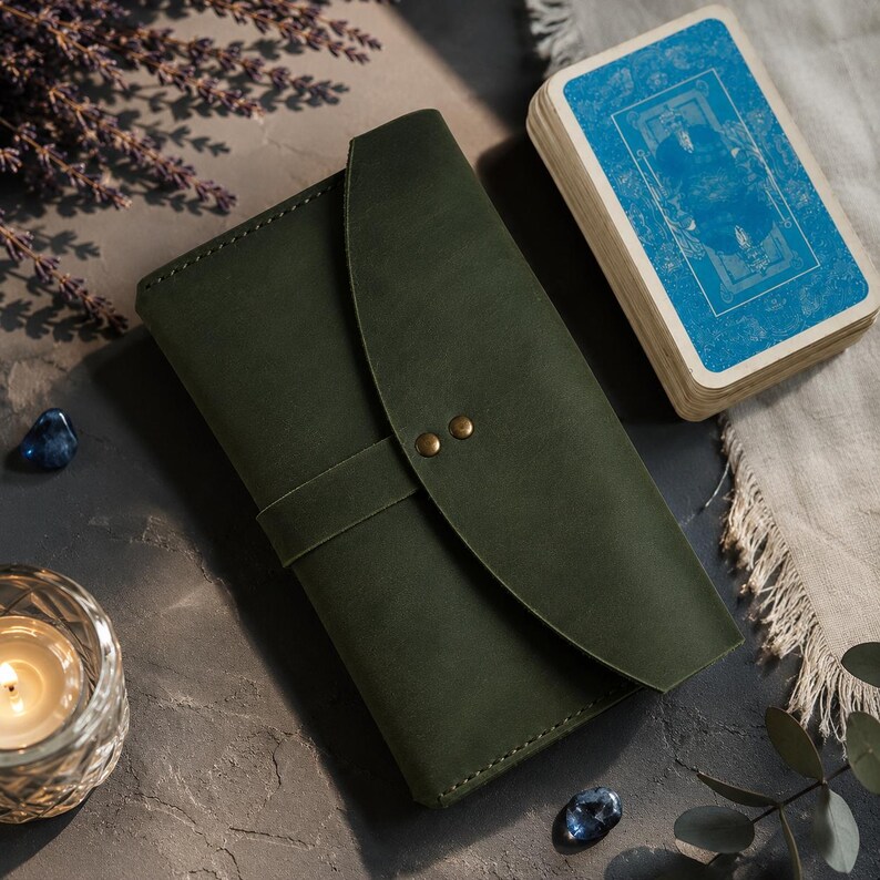 May include: A dark olive green leather journal cover featuring a curved flap closure secured by two small brass-colored metal rivets. The rectangular cover has visible stitching along the edges and a horizontal strap that holds the flap in place.