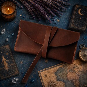 May include: A rectangular brown leather pouch featuring a wrap-around leather tie closure. The pouch has a smooth, matte finish with visible stitching along the edges and a long, thin leather strap that secures the flap in a horizontal orientation.