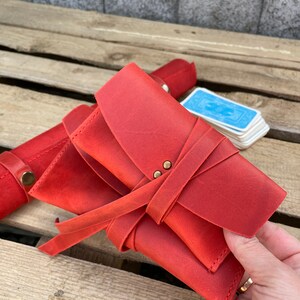 May include: A red leather card case with a tie closure and brass accents. The case is folded and held in a hand.