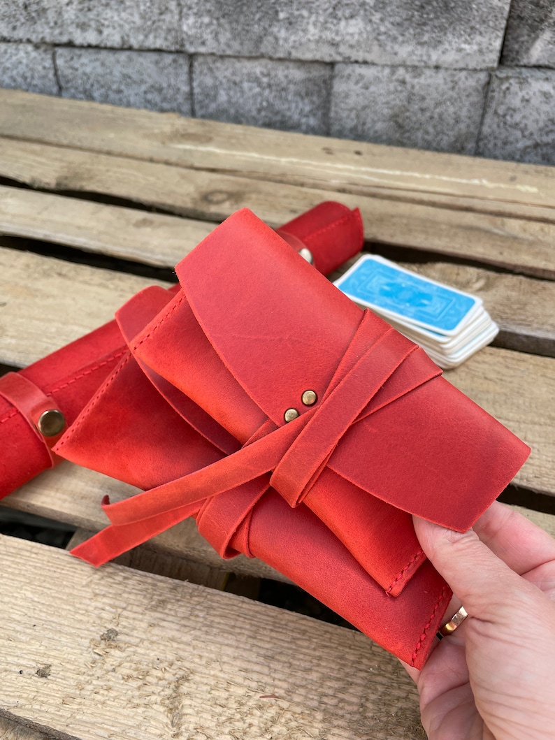May include: A red leather card case with a tie closure and brass accents. The case is folded and held in a hand.