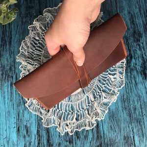 May include: A rectangular brown leather case with a smooth finish and a curved flap closure. The case is secured by a thin leather strap that wraps around the center, featuring visible stitching along the edges and a simple, functional design.