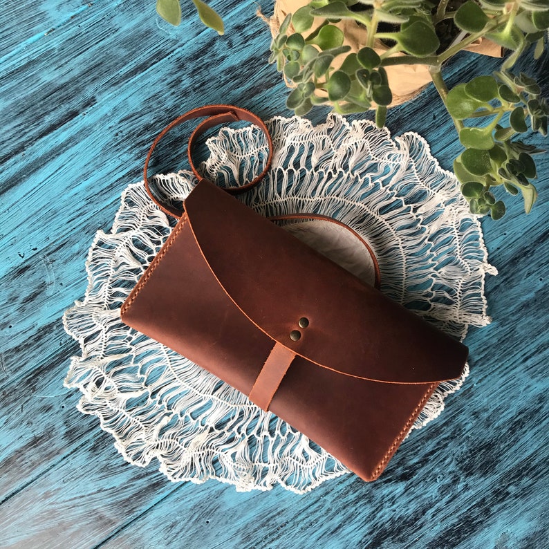 May include: A dark brown leather clutch bag with a curved flap closure, secured by a thin leather strap and two small metal rivets. The bag features a long, thin leather shoulder strap and visible stitching along the edges of the main body.