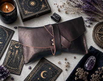 May include: A dark brown leather pouch with a wrap-around closure secured by two metallic rivets. The pouch features a textured surface, visible stitching along the edges, and an asymmetrical flap design that folds over the main compartment.