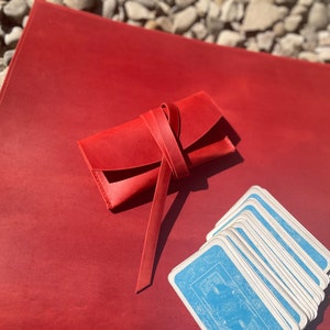 May include: A red leather card case with a tied closure, sitting on a red leather surface with a deck of playing cards.