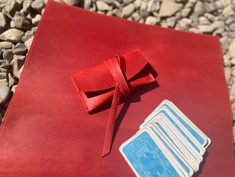 May include: A red leather card case with a tied closure, sitting on a red leather surface with a deck of playing cards.