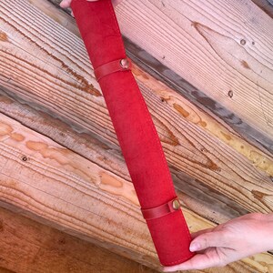 May include: A red leather pencil roll with two brass clasps. The roll is rolled up and held by a hand.