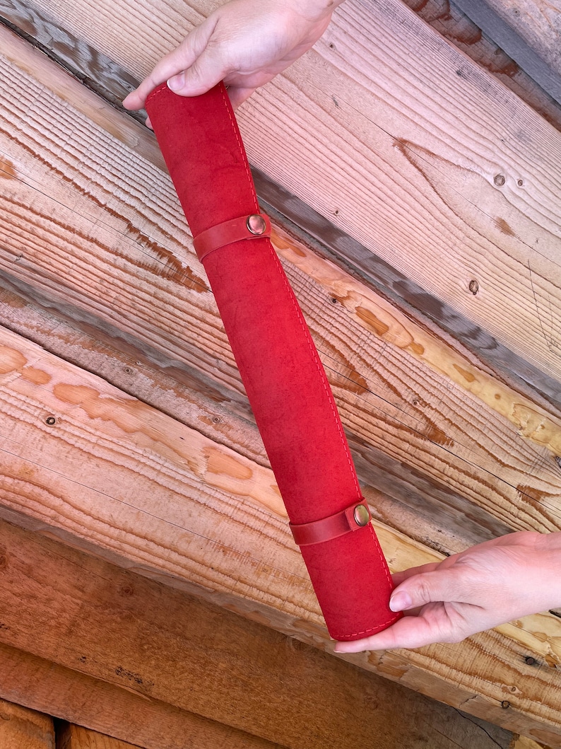 May include: A red leather pencil roll with two brass clasps. The roll is rolled up and held by a hand.