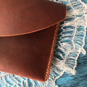 May include: A brown leather pouch featuring a smooth, matte surface and a curved flap closure. The edges of the pouch are secured with visible, thick tan stitching that runs along the side, highlighting the construction of the leather accessory.