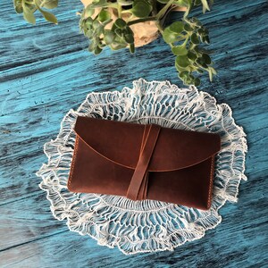 May include: A rectangular brown leather wallet featuring a smooth matte finish and a curved flap closure. The wallet is secured by a thin leather strap that wraps around the center, holding the flap firmly in place against the main body of the item.