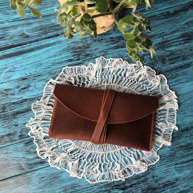 May include: A rectangular brown leather wallet featuring a smooth matte finish and a curved flap closure. The wallet is secured by a thin leather strap that wraps around the center, holding the flap firmly in place against the main body of the item.