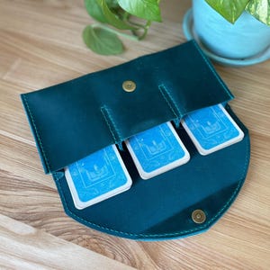 Könnte beinhalten: Tarotkartenhalter aus tealfarbenem Leder mit drei Kartenfächern, gesichert durch einen Druckknopfverschluss. Der Halter enthält drei Decks mit blauen Tarotkarten. Die Nähte sind in einem helleren Teal gehalten. Der Halter befindet sich auf einer Holzoberfläche.