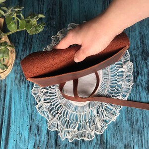 May include: A brown leather pouch featuring a smooth exterior and a textured, unfinished interior surface. The pouch includes a long, thin leather strap attached to the side, allowing for closure or carrying, with visible grain details on the leather.