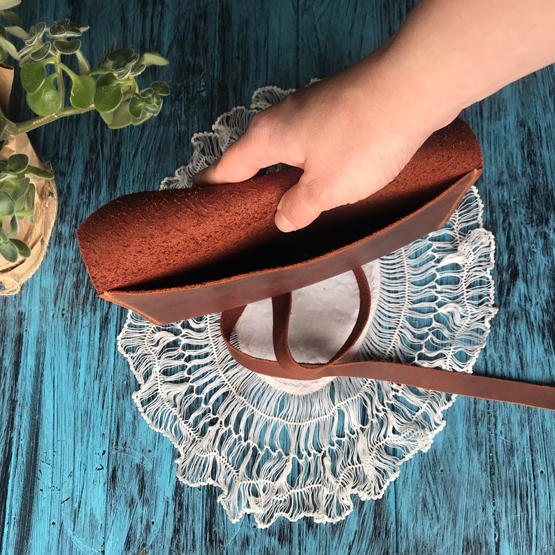 May include: A brown leather pouch featuring a smooth exterior and a textured, unfinished interior surface. The pouch includes a long, thin leather strap attached to the side, allowing for closure or carrying, with visible grain details on the leather.