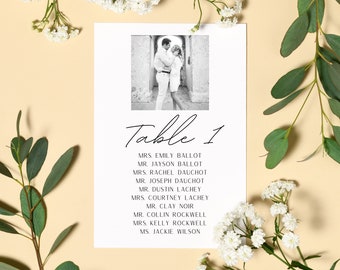 May include: A wedding table seating card with a black and white photo of a couple. The card reads "Table 1" and lists names below. White flowers and green leaves surround the card on a light yellow background.