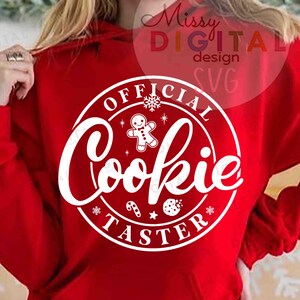 May include: A red hooded sweatshirt with a white circular graphic that reads "OFFICIAL Cookie TASTER". The graphic includes a gingerbread man, snowflakes, and other festive elements. The sweatshirt is a solid color.