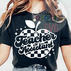 May include: Black t-shirt featuring a white and black checkered apple design with the words "Teacher Assistant" in a retro font. The design includes white star accents. A casual, comfortable top.
