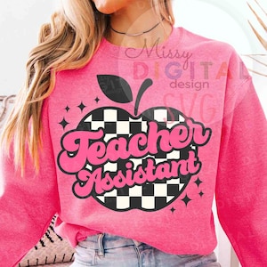 May include: A pink sweatshirt with a graphic of an apple with a black and white checkered pattern. The words "Teacher Assistant" are in pink, retro-style lettering. The design includes black stars and a black apple stem.