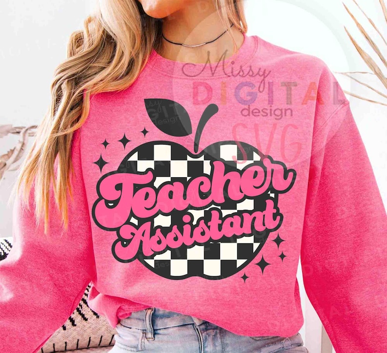 May include: A pink sweatshirt with a graphic of an apple with a black and white checkered pattern. The words "Teacher Assistant" are in pink, retro-style lettering. The design includes black stars and a black apple stem.
