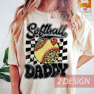 Op de afbeelding: Een wit T-shirt met een zwart-wit geruite achtergrond, een softball met een luipaardprint en de tekst "Softball Daddy" in een verweerd lettertype.